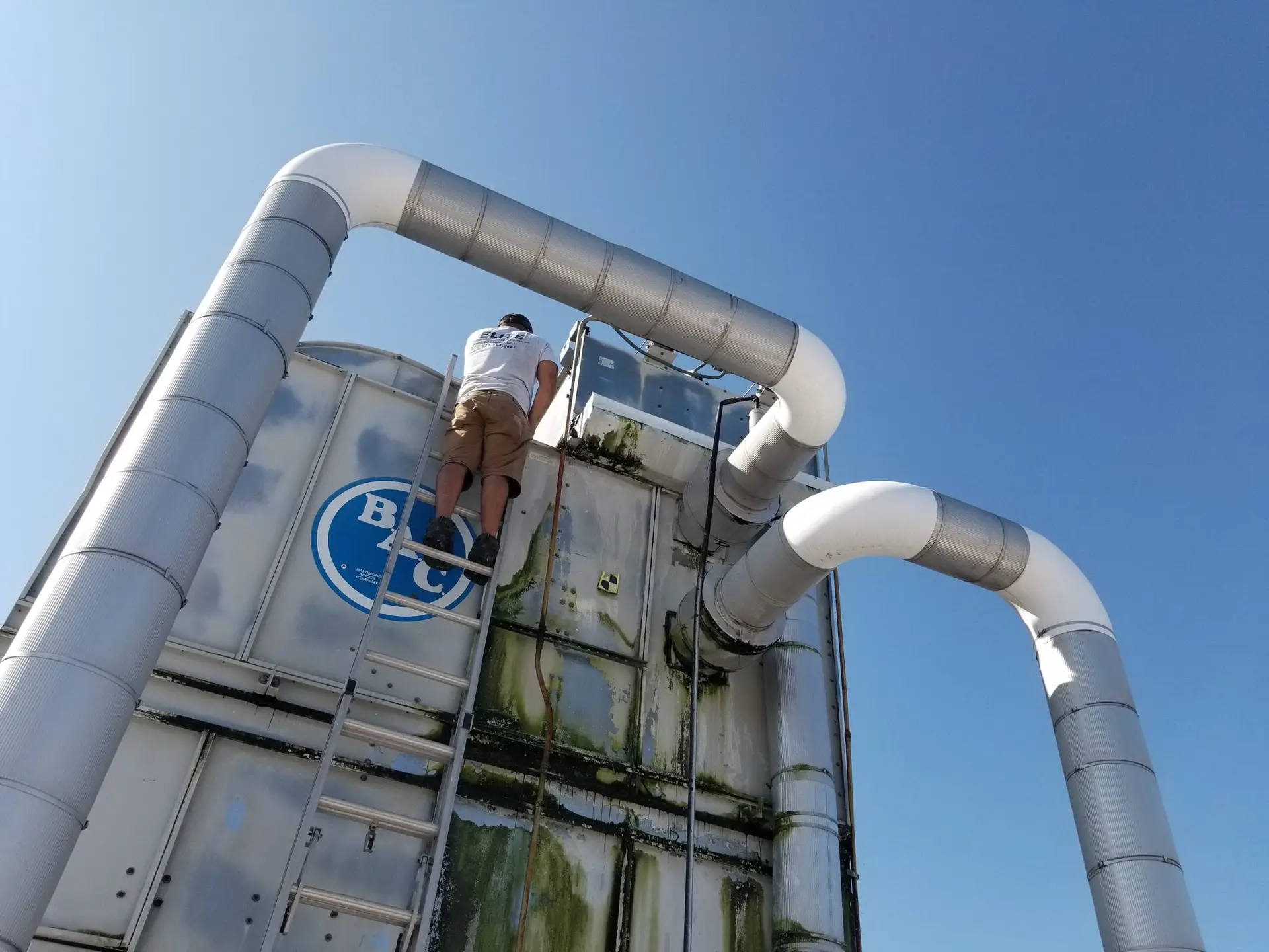 A person in a white shirt and shorts climbs a tall industrial structure with large, curved pipes against a clear blue sky. The structure has green stains and a blue logo with white text. The person is on a metal ladder near the top of the structure.