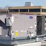 A rooftop HVAC unit with a Carrier logo is mounted alongside solar panels on a building. In the background, there is a multi-story office building with reflective windows. The surrounding area has bare trees and a clear sky, showcasing why Commercial HVAC repair services are crucial here.