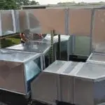 A complex network of reflective, metallic air ducts on a rooftop showcases straight and angular sections. Connected with visible sealing at the joints, a person in a striped shirt focuses on Commercial HVAC repair. Dense green trees provide a scenic backdrop to this precise operation.