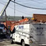 A work crew focuses on commercial HVAC repair, operating on utility lines using a crane and a bucket truck in a narrow alley. Orange cones mark the area. A white van with Elite Heating and Air Conditioning written on it is parked nearby, showcasing their expertise among the best air conditioning repair companies.