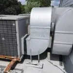 A commercial rooftop HVAC system with large metal ducts and a weathered outdoor unit awaits repair. The metal casing shows rust and wear. The ducts connect to the building’s side, with greenery and trees in the background under a partly cloudy sky.