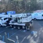 A parking lot featuring a large crane truck labeled NATIONAL CRANE occupying two spaces, including a handicapped spot. Nearby, two white service vans are parked with ELITE branding. The ground is marked with blue handicapped symbols, and trees border the lot.