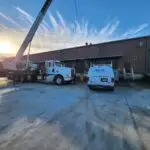 A construction scene with a white crane truck and a white van parked on a concrete yard in front of a brick warehouse. Workers are visible on the roof of the building, with a clear blue sky and the sun partially obscured by clouds in the background.