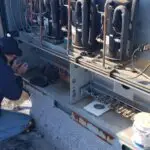A technician in a dark hoodie and cap kneels, inspecting a large commercial HVAC unit. The open panel reveals pipes, wires, and components. Nearby are tools, a drill, gloves, and a paint can. The technician's shirt reads Elite Heating & Air with a phone number on the back.