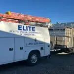 A white service truck labeled Elite Heating and Air Conditioning Inc. of Cherry Hill is parked on gravel. It has a phone number displayed. Three orange and red ladders are secured on top. A trailer hitched behind carries stacked industrial units. A clear blue sky is overhead.