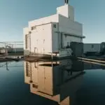 A person stands atop a white rooftop structure, reflected in a large puddle below. The clear blue sky creates a serene atmosphere, while industrial elements like pipes and vents evoke thoughts of the best HVAC companies nearby, adding contrast to the peaceful reflection in the water.