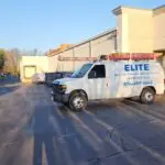 A white van with ELITE Heating and Air Conditioning 856-489-8664 printed on the side is parked outside a commercial building. A ladder is mounted on its roof, likely for a rooftop repair. The building has beige walls and a green awning. It's sunny, with long shadows indicating late afternoon.
