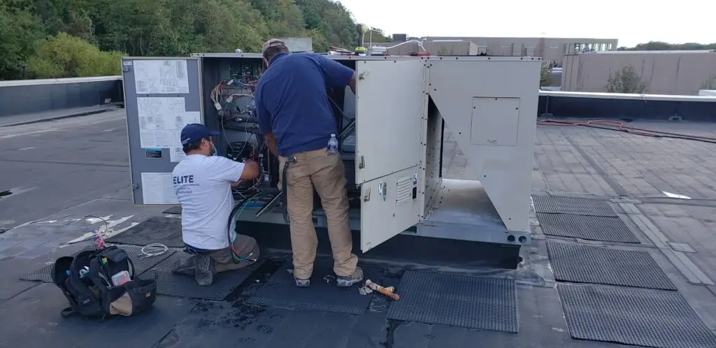 Two technicians work on a rooftop HVAC unit. One kneels, wearing a white shirt and dark shorts, while the other stands in a blue shirt and tan pants. Tools and a bag are nearby, signaling an air conditioning repair in progress. Trees and a commercial building are visible in the background.