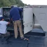 Two technicians work on a rooftop HVAC unit. One kneels, wearing a white shirt and dark shorts, while the other stands in a blue shirt and tan pants. Tools and a bag are nearby, signaling an air conditioning repair in progress. Trees and a commercial building are visible in the background.
