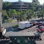 A commercial flatbed truck with a man seated in the driver's cabin is parked in a lot. Behind it, a white van labeled Elite is parked next to red and silver cars. Trees line the background, partially obscuring an office building with large windows, hinting at Moorestown's industrial landscape.