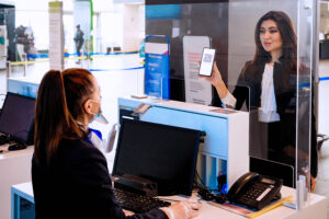 A woman at an airport check-in counter in South Jersey shows a QR code on her phone to a staff member behind a plexiglass barrier. The staff member wears a mask and gloves, seated at a computer. The setting is bright and modern, with barriers and signs in the background.