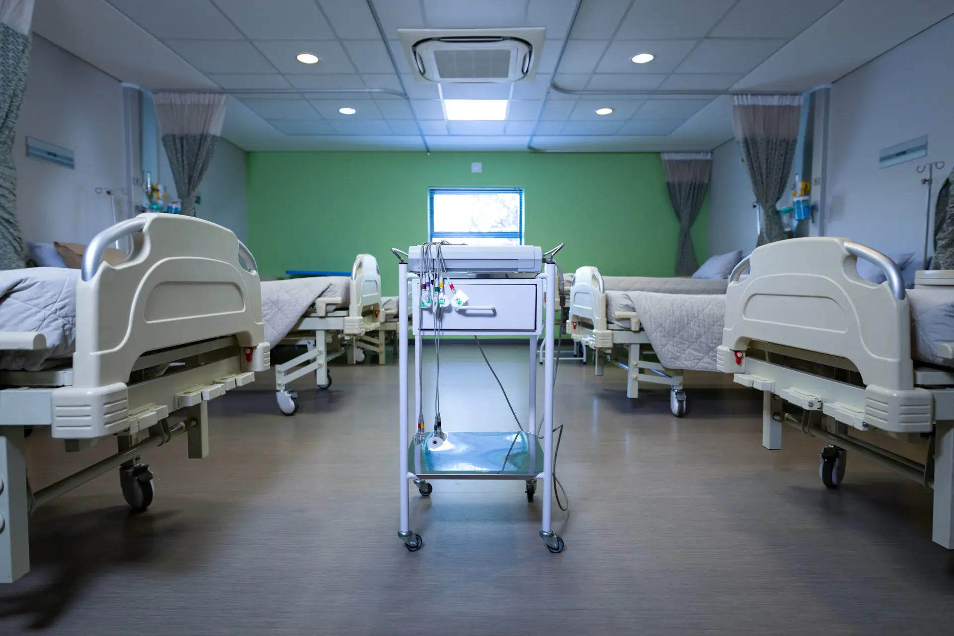 A hospital room in South Jersey with four empty beds, privacy curtains, medical equipment on a cart in the center, and hospital air conditioning ensuring comfort under fluorescent lighting.