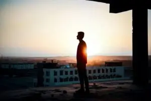 Silhouette of a person standing in a building under construction, facing a sunset. They stand on the concrete floor, contemplating ways to improve building performance. The sky is a gradient of warm colors with the sun setting beyond distant rooftops and flat terrain, evoking tranquility and reflection.