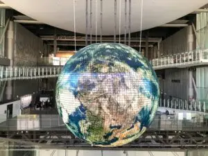 A large digital globe display, designed with Elementor, is suspended from the ceiling in a modern exhibition space. The globe shows Earths continents in vivid detail. The room features high ceilings, walkways, and visitors observing the installation from various platforms. Light filters in from windows above.