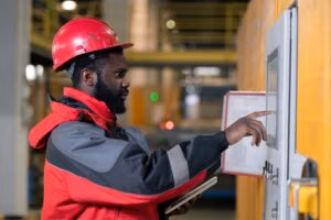 A man in a red hard hat and red-and-gray work jacket uses a touchscreen control panel in an industrial setting, intent on providing preventative maintenance.