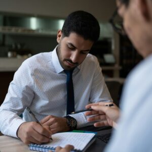 Two men in dress shirts sit at a table; one writes notes in a notebook while the other gestures with a pen during a discussion about the hidden toll of humidity in commercial buildings.