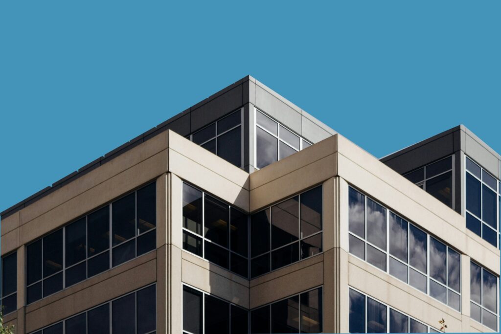 Corner view of a modern office building with large glass windows and beige exterior panels, illustrating how humidity can take a hidden toll on commercial buildings, set against a clear blue sky.