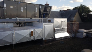 Commercial HVAC units and insulated air ducts are installed on a flat rooftop beside a brick chimney, capturing unusual moments under the partly cloudy 2025 sky.
