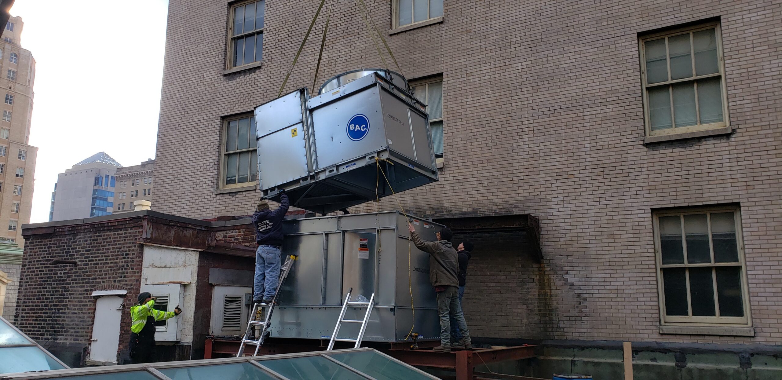 A large Commercial HVAC unit is lifted by a crane onto a rooftop in 2025 as three workers guide it into place next to a brick building, capturing one of those unusual moments on the job site.