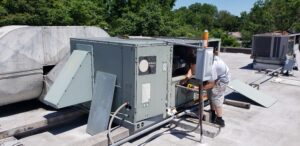 A technician is servicing a large Commercial HVAC unit on a flat commercial building under a clear 2025 sky, surrounded by trees—an unusual moment of calm in the busy world of rooftop repairs.