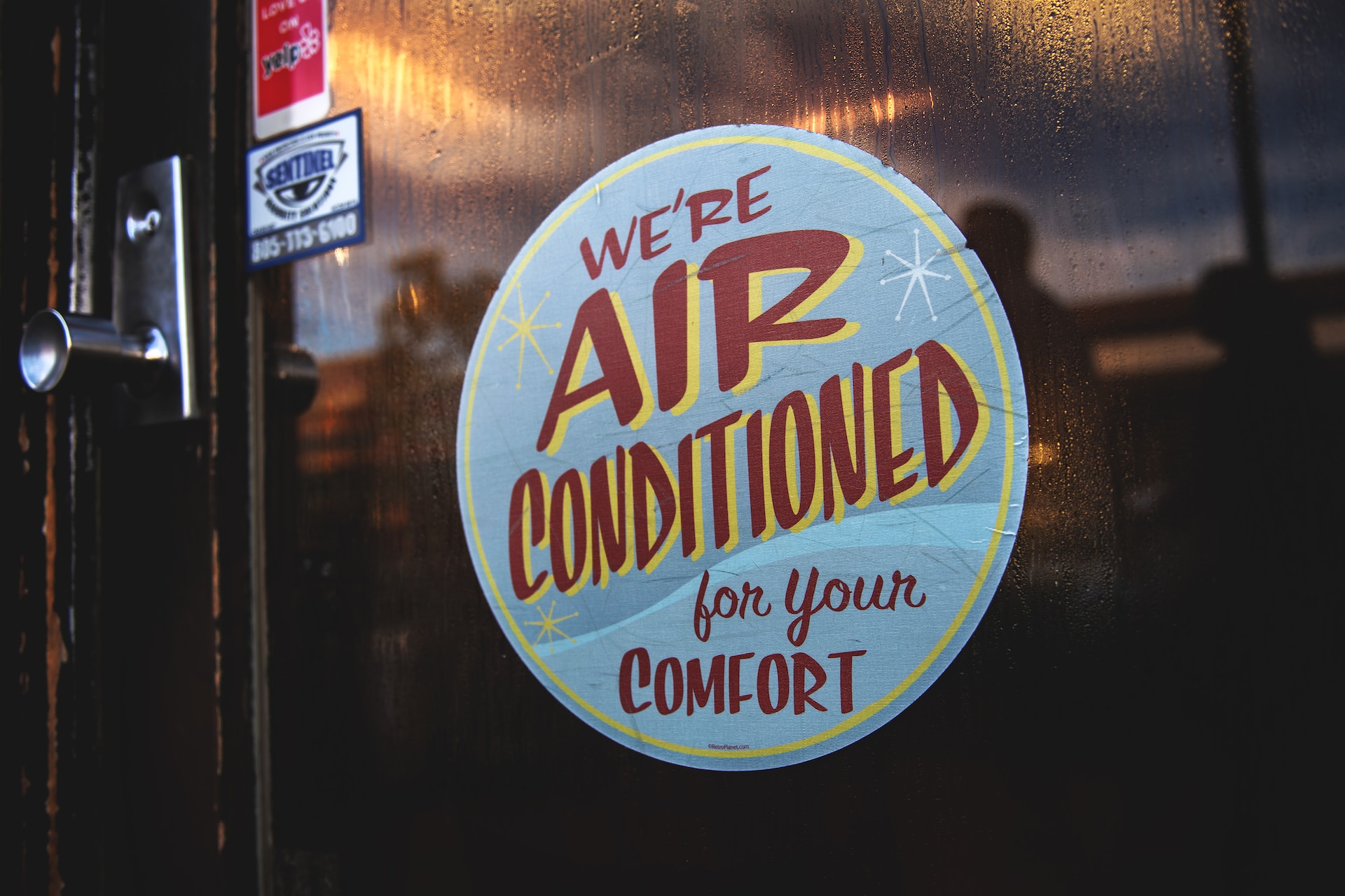A round sign on a glass door reads “We’re air conditioned for your comfort” in bold, colorful letters—a nod to commercial HVAC and those unusual moments of relief, even as we head into 2025.