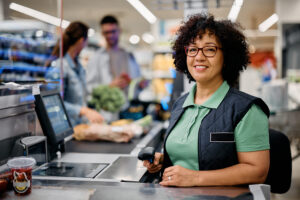 Smiling supermarket cashier holds a barcode scanner at her register, with customers and groceries visible in the background—capturing one of those unusual commercial HVAC moments you might notice in 2025.