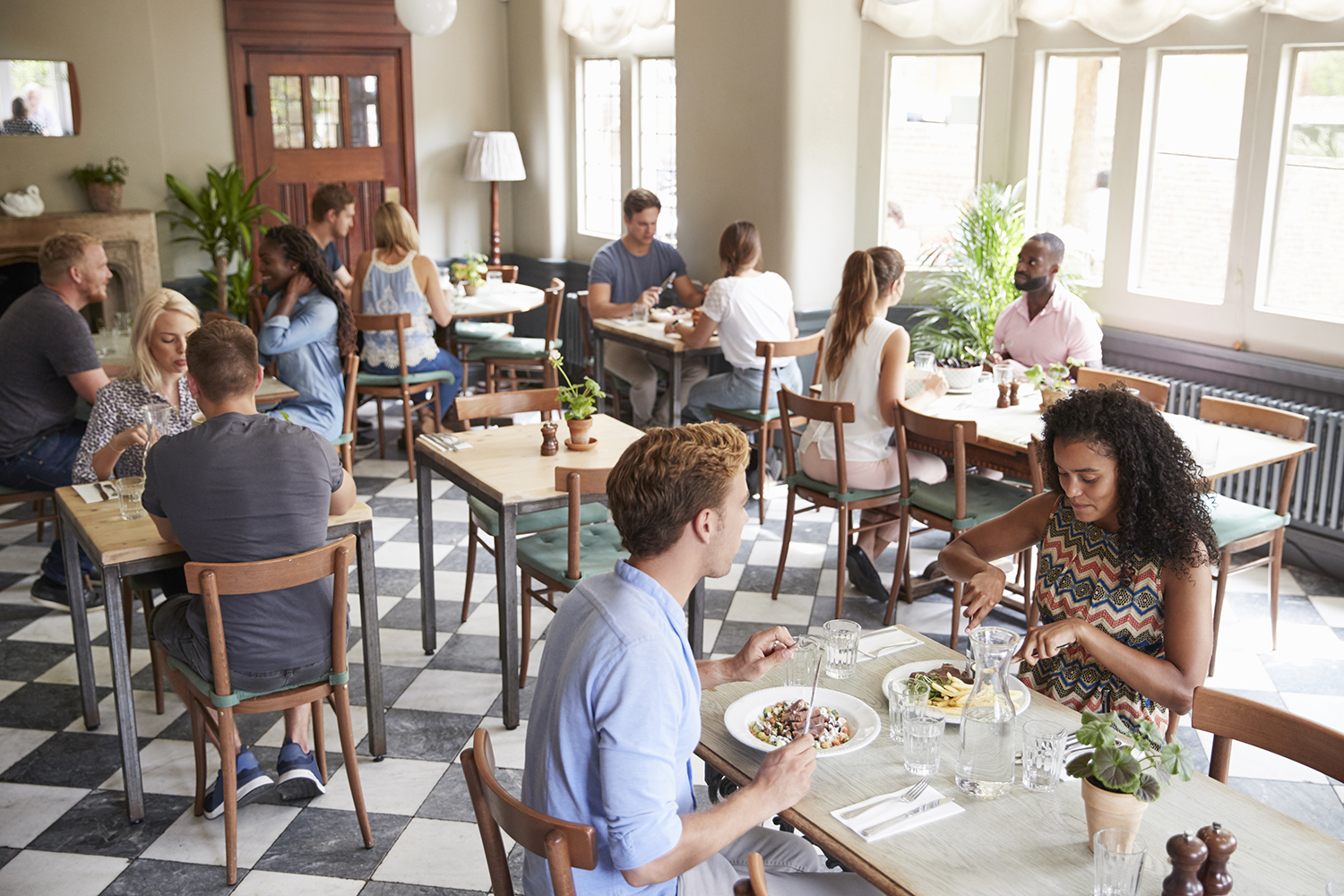 People sit at tables eating and talking in a bright, casual restaurant with checkered floors and large windows letting in natural light, while enjoying the comfort of a Commercial HVAC system. Unusual moments make 2025 dining more memorable.