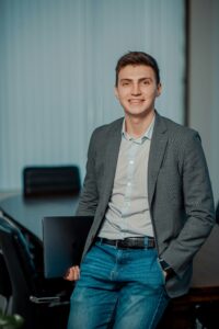 A man in a gray blazer and jeans stands in an office, holding a closed laptop and smiling at the camera—capturing one of those unusual moments in a busy commercial HVAC firm.