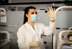 A woman in a lab coat, gloves, and a surgical mask holds up a small black object in a clinical or laboratory setting, capturing one of those unusual moments reminiscent of behind-the-scenes tests for cutting-edge commercial HVAC systems in 2025.