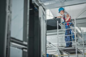 A construction worker in safety gear stands on scaffolding, inspecting a commercial HVAC duct inside an industrial building under construction—capturing one of those unusual moments found on the job site in 2025.
