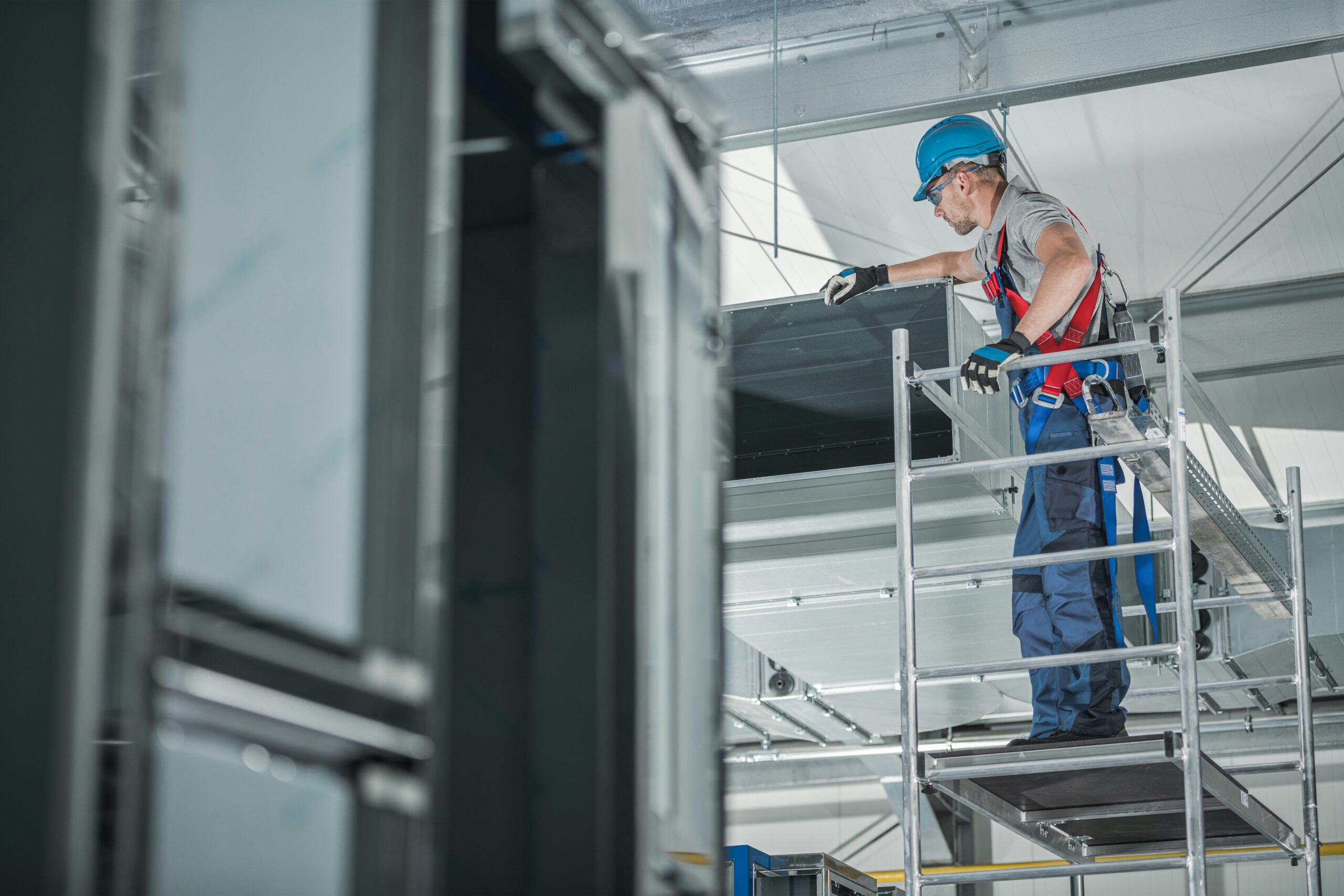 A construction worker in safety gear stands on scaffolding, inspecting a commercial HVAC duct inside an industrial building under construction—capturing one of those unusual moments found on the job site in 2025.