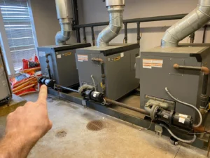 A person points toward a row of Commercial HVAC units connected by metal pipes inside a maintenance room, capturing one of those unusual moments behind the scenes.
