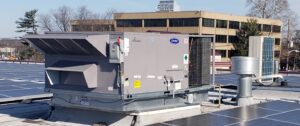 A large commercial HVAC unit sits next to solar panels and other equipment atop a rooftop, with office buildings and trees in the background—capturing one of 2025’s unusual moments in sustainable design.