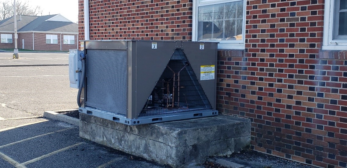 A large Commercial HVAC unit is installed on a concrete platform outside a brick building, near a window and an empty parking lot, ready to deliver reliable climate control for 2025.