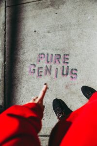A person in a red sleeve points to the words PURE GENIUS stenciled in pink on a concrete sidewalk, capturing one of those unusual moments in 2025, with their black shoes visible.