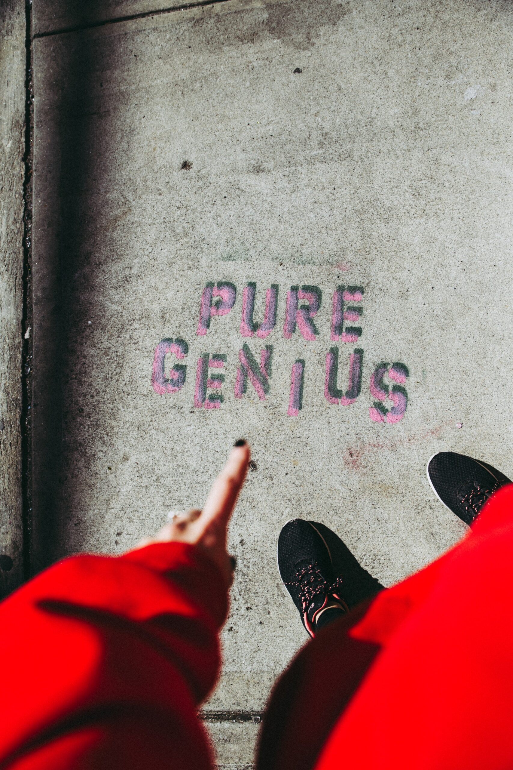 A person in a red sleeve points to the words PURE GENIUS stenciled in pink on a concrete sidewalk, capturing one of those unusual moments in 2025, with their black shoes visible.