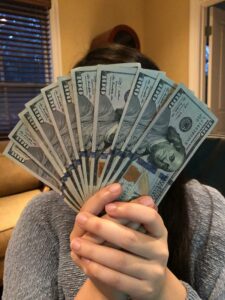 A person holds a fan of eight $100 bills, partially covering their face, indoors near a window and beige furniture, capturing one of those Unusual Moments that 2025 is sure to bring.