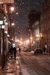 Snow falls on a dimly lit city street at night, where warm streetlights reveal unusual moments—like the gentle hum of a commercial HVAC unit blending with quiet footsteps as people walk past parked cars.