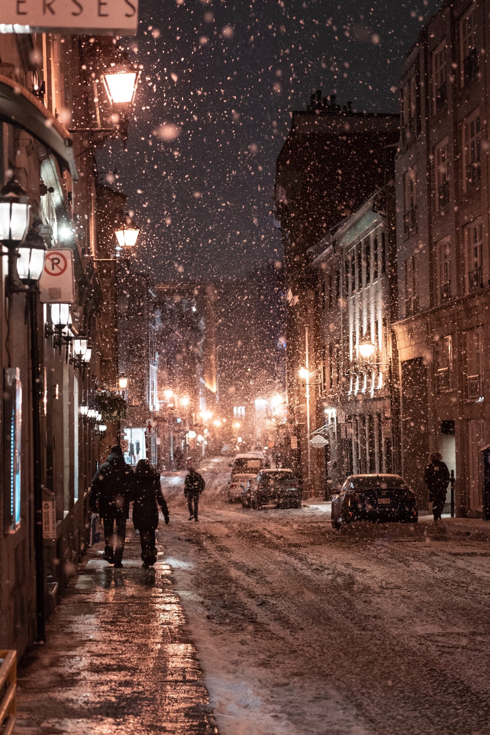 Snow falls on a dimly lit city street at night, where warm streetlights reveal unusual moments—like the gentle hum of a commercial HVAC unit blending with quiet footsteps as people walk past parked cars.