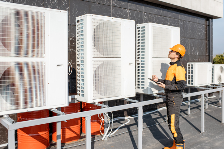 A technician in a hard hat and uniform inspects multiple outdoor Commercial HVAC units mounted on a metal platform against a building wall, prepared for any unusual moments in 2025.