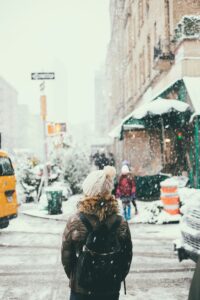 Person in winter clothing with a backpack stands on a snow-covered city street during Unusual Moments of 2025; snow is falling and other people, cars, and buildings—some fitted with commercial HVAC units—are visible in the background.
