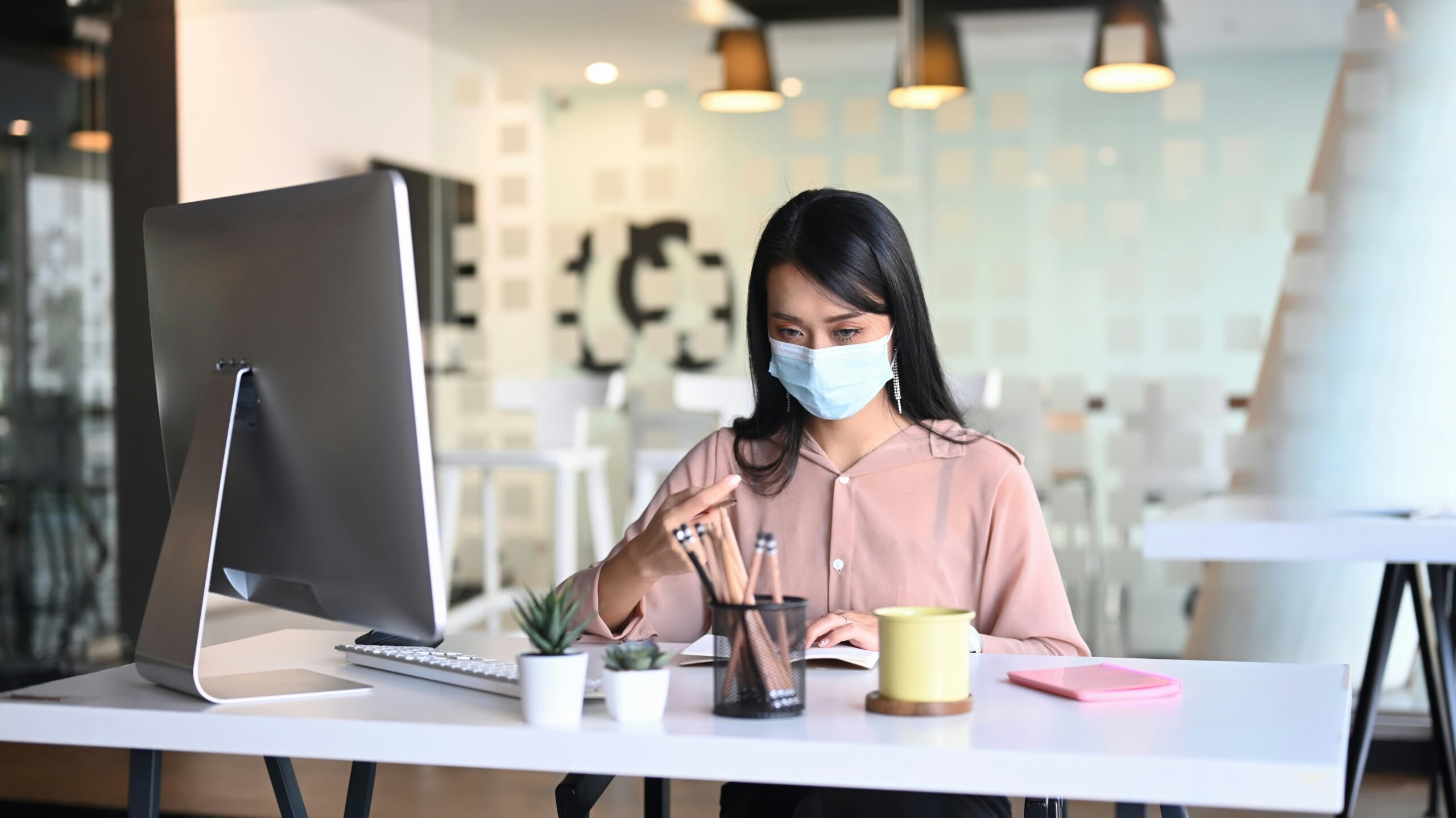 A woman wearing a face mask works at a desk with a computer, stationery, and a coffee mug in a modern office setting.