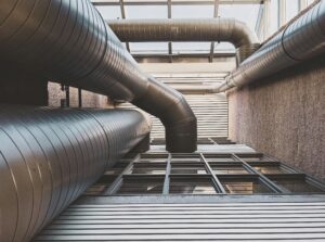 Large industrial air ducts run along the ceiling and walls of a building interior, with metal panels and glass windows visible below.