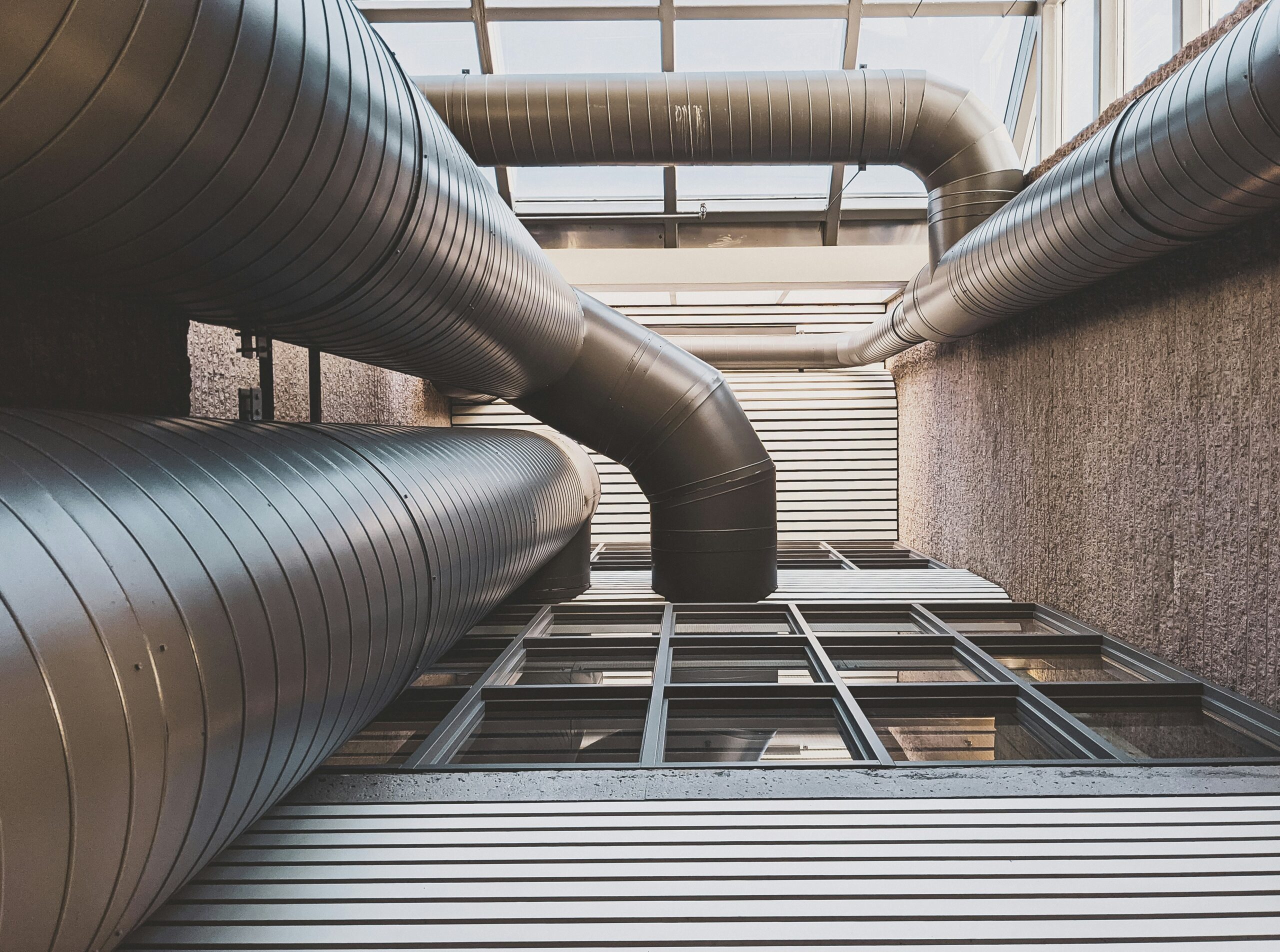 Large industrial air ducts run along the ceiling and walls of a building interior, with metal panels and glass windows visible below.