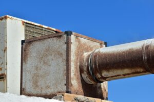 A close-up of a rusty metal ventilation unit and duct against a clear blue sky.