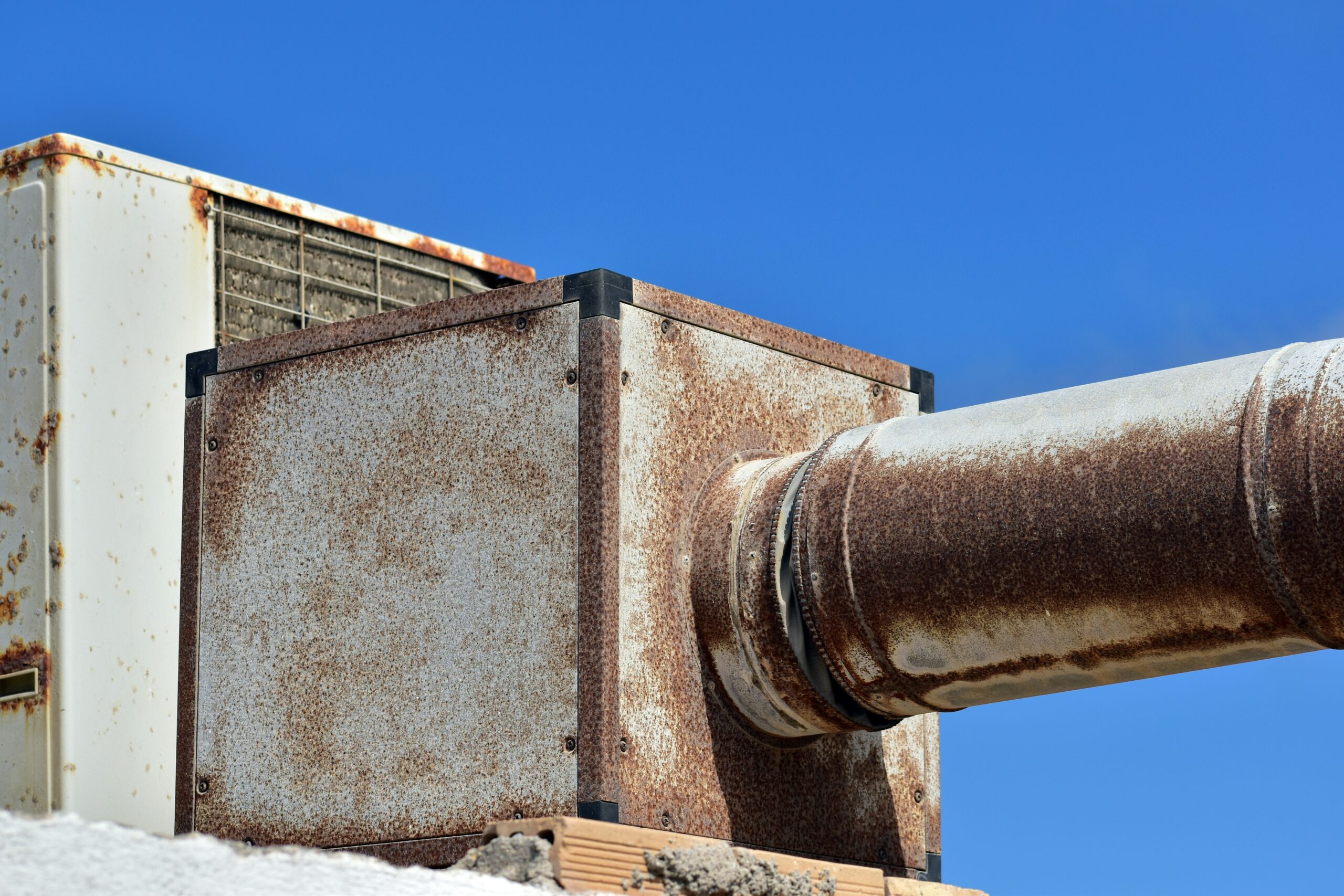 A close-up of a rusty metal ventilation unit and duct against a clear blue sky.