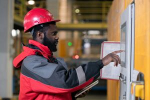 A worker in a red hard hat and jacket operates a touchscreen control panel in an industrial setting, holding a clipboard under one arm.