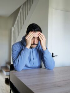 A man in a blue hoodie sits at a table with his head in his hands, appearing stressed or overwhelmed. A staircase and door are visible in the background.
