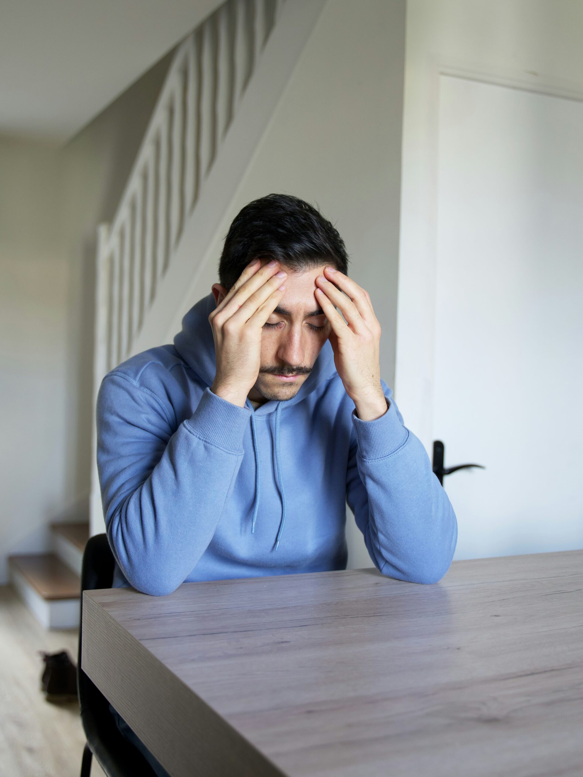 A man in a blue hoodie sits at a table with his head in his hands, appearing stressed or overwhelmed. A staircase and door are visible in the background.