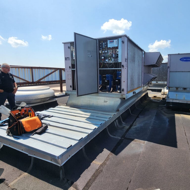 A rooftop HVAC unit with its control panel open, tools and a tool bag nearby, and a person standing to the side under a clear sky.